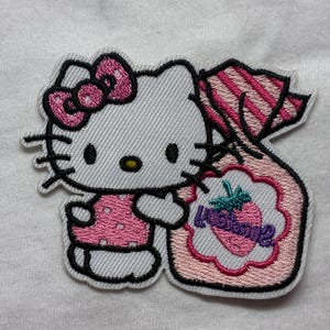 Peut inclure: Écusson brodé représentant un chat blanc avec un nœud et une robe roses, tenant un sac rose avec un motif de fraise et le mot "In Store". L'écusson a des contours noirs et est sur fond blanc.