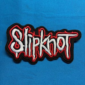 May include: An embroidered patch featuring the word Slipknot in a stylized white font with a red outline, all set against a black background. The lettering has a jagged, distressed appearance with sharp edges and a distinct, bold aesthetic.