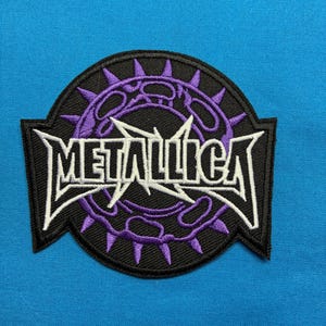 May include: A circular embroidered patch featuring the white Metallica logo text centered over a purple spiked crown design. The patch has a black background with a thick black border and extends into two side points on the left and right edges.