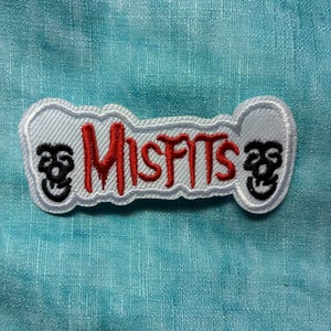 May include: Embroidered patch featuring the word "Misfits" in red, stylized lettering. The patch has a white border and includes two skull graphics. The background is a textured, light blue fabric.