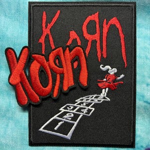 Könnte beinhalten: Schwarzer rechteckiger Aufnäher mit dem Bandnamen "Korn" in roter, stilisierter Schrift. Ein Hüpfspiel ist in Weiß dargestellt, mit einer kleinen Figur in einem roten Kleid. Der Aufnäher befindet sich auf einem blau-türkisfarbenen Hintergrund.