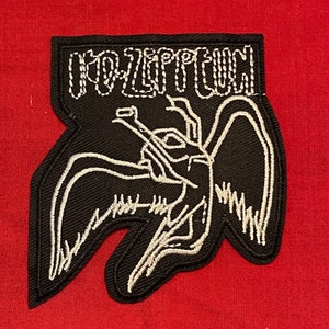 Puede incluir: Parche negro con texto bordado blanco "LED ZEPPELIN" sobre una ilustración blanca de una figura alada. El parche tiene una forma única y está sobre un fondo rojo.