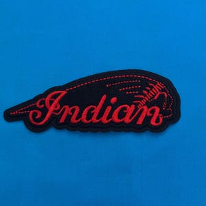 Pode incluir: Patch bordado preto e vermelho com a palavra "Indian" em escrita estilizada. O patch tem um fundo preto e uma borda vermelha, com um design vermelho no lado direito. O patch está sobre um fundo azul brilhante.