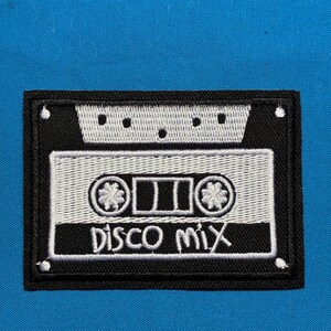 May include: A rectangular embroidered patch featuring a black and white cassette tape design on a bright blue fabric background. The patch displays the text "Disco Mix" in white embroidery, with a detailed reel and tape housing graphic.