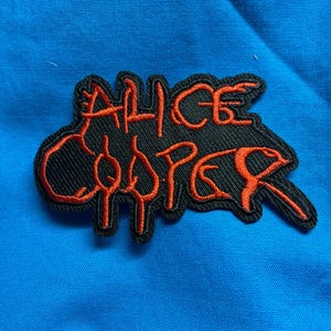 May include: An embroidered patch featuring the text Alice Cooper in a stylized, dripping orange font on a black background. The lettering has a distinct, jagged appearance with elongated strokes, creating a bold contrast against the dark fabric base.