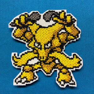 May include: A cross-stitch patch featuring a pixelated, yellow, bipedal creature with dark brown accents, pointed ears, and two long, flowing appendages extending from its sides, all outlined in white against a solid blue background.