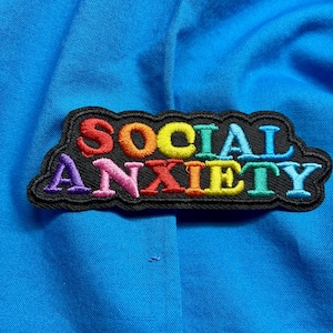 May include: A rectangular embroidered patch with a black background and a scalloped edge. The text SOCIAL ANXIETY is stitched in a rainbow gradient of colours, featuring red, orange, yellow, green, blue, indigo, and violet thread on blue fabric.