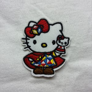 Peut inclure: Écusson brodé représentant un personnage de chat blanc avec un nœud rouge, tenant une version miniature de lui-même. Le chat porte un manteau rouge avec un motif géométrique coloré et des chaussures marron. L'écusson a une bordure blanche.