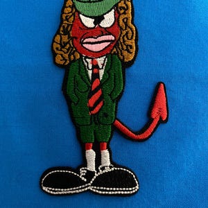 Peut inclure: Un patch brodé représentant un diable de dessin animé rouge portant un chapeau vert, une veste verte, une cravate rouge et des chaussures noires. Le diable a une queue rouge et des cheveux bruns bouclés.