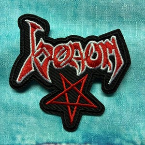 May include: Embroidered patch featuring the word "Venom" in red, outlined in white, on a black background. A red pentagram is below the word. The patch is likely for a jacket or bag.
