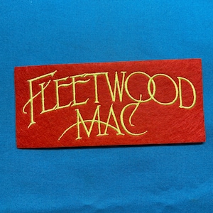 May include: A rectangular red felt patch featuring the band name Fleetwood Mac embroidered in yellow thread. The text is written in a stylized script font with the word Fleetwood on the top line and Mac on the bottom line, centered.