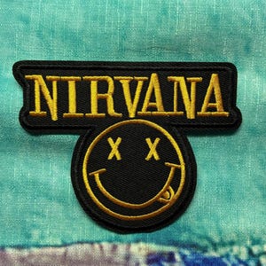 May include: Black embroidered patch with the word "NIRVANA" in gold lettering above a black smiley face with crossed-out eyes and a tongue, outlined in gold. The patch is on a teal and blue fabric background.