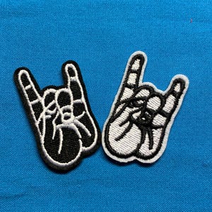 Peut inclure: Deux écussons brodés représentant une main faisant le signe du rock and roll. Un écusson présente une main noire avec des contours et des détails blancs, tandis que l'autre présente une main blanche avec des contours et des détails noirs sur un tissu bleu.