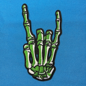 Könnte beinhalten: Ein gestickter Aufnäher, der eine skelettierte Hand zeigt, die ein Rock-and-Roll-Zeichen macht. Die Knochen sind mit leuchtend grünem und weißem Garn und einem dicken schwarzen Rand auf einem einfarbigen blauen Stoffhintergrund dargestellt.