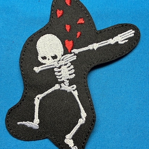 May include: An embroidered patch featuring a white skeleton performing a dabbing pose against a black background. The skeleton is surrounded by several small, red embroidered hearts floating above its head, all contained within a black border.