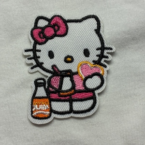 以下が含まれることがあります： ピンクのリボンをつけた白い漫画の猫が、ハート型のクッキーとボトルを持っている刺繍パッチ。猫はピンクの服を着ています。パッチは黒いアウトラインで、白い生地の背景に配置されています。