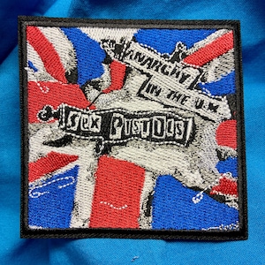 Op de afbeelding: Een vierkante geborduurde patch met een Union Jack vlagachtergrond in rood, wit en blauw. De patch toont de tekst "ANARCHY IN THE U.K." en "SEX PISTOLS" in een vet, verweerd zwart lettertype met een dikke zwarte rand rondom de rand.