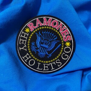 May include: A round, embroidered patch with the text "RAMONES" in pink, surrounded by yellow stars. The patch features a blue eagle design and the phrases "HEY HO LET'S GO" in white. The patch is on a blue fabric background.