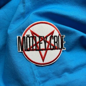 Puede incluir: Un parche bordado circular que presenta un pentagrama rojo sobre un fondo blanco con el texto negro MÖTLEY CRÜE superpuesto en el centro. El parche tiene un borde rojo cosido y está colocado sobre una superficie de tela azul brillante.