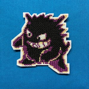 May include: An embroidered patch featuring a stylized dark purple and black creature with a wide, toothy grin and pointed ears. The design is outlined in white thread, showcasing a jagged, spiky silhouette and detailed stitching throughout.