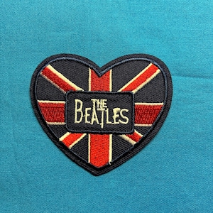 May include: A heart-shaped embroidered patch featuring a Union Jack flag design in red, white, and black. Centered on the patch is a black rectangular border containing the white embroidered text that reads THE BEATLES.