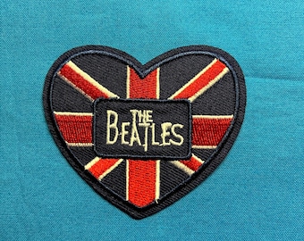 May include: A heart-shaped embroidered patch featuring a Union Jack flag design in red, white, and black. Centered on the patch is a black rectangular border containing the white embroidered text that reads THE BEATLES.