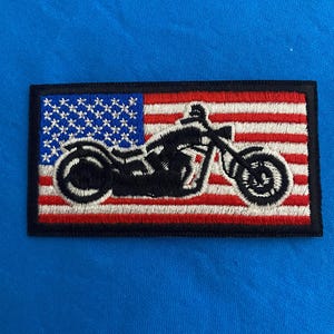 May include: An embroidered patch featuring a black motorcycle silhouette in front of an American flag. The flag is red, white, and blue with white stars on a blue field.