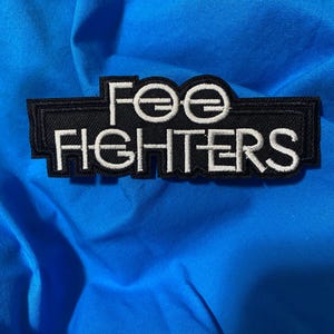 Könnte beinhalten: Ein schwarz-weiß gestickter Aufnäher mit dem Text "Foo Fighters" auf blauem Stoff. Der Aufnäher hat eine rechteckige Form mit dem Namen der Band in einer stilisierten Schriftart. Der Hintergrundstoff ist einfarbig hellblau.