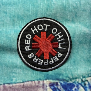 Könnte beinhalten: Ein runder Aufnäher mit den Worten "RED HOT CHILI PEPPERS" in weißer Schrift am Rand. In der Mitte befindet sich ein rotes sternchenartiges Design. Der Aufnäher befindet sich auf einem blau-violetten Stoffhintergrund.
