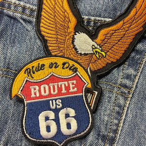 Puede incluir: Parche bordado con un águila dorada con las alas extendidas sobre un letrero de la Ruta 66. El letrero es rojo, blanco y azul con las palabras "ROUTE US 66" y el número "66". El parche está sobre un fondo de mezclilla.
