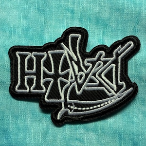 May include: Black and white embroidered patch with the word "HAZEL" in a stylized font. The patch has a black border and is set against a teal and purple fabric background.