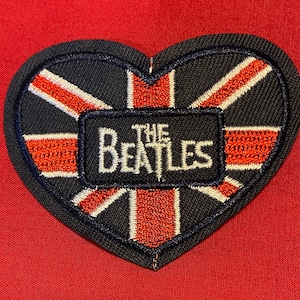 May include: A heart-shaped patch featuring a Union Jack design with the words "The Beatles" in white lettering on a black background.