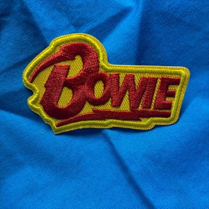 Könnte beinhalten: Ein gestickter Aufnäher mit dem Wort Bowie in rotem Garn und einem Blitz-Akzent, platziert auf einem gelben Hintergrund mit gelbem Rand. Der Aufnäher liegt auf einer leuchtend blauen Stoffoberfläche mit sichtbaren Falten und Textur.