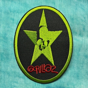 May include: Embroidered patch featuring a green star design within a black oval, outlined in green. The word "Gorillaz" is written in red script below the star. The patch is likely for clothing or accessories.