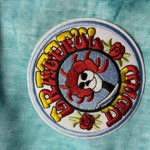 Könnte beinhalten: Runder, gestickter Aufnäher mit den Worten "Grateful Dead" in Gelb und Rot. Das Design zeigt einen Totenkopf mit roter Blume, vor blauem Hintergrund und weißem Rand.