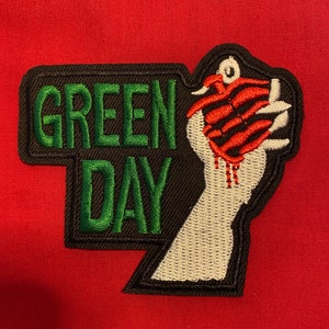 Puede incluir: Parche bordado con las palabras "GREEN DAY" en letras verdes sobre un fondo negro. El parche incluye una mano blanca agarrando un diseño rojo en forma de garra, con goteo rojo. El parche está sobre un fondo rojo.
