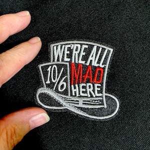 May include: Embroidered patch featuring a Mad Hatter hat design with the text "WE'RE ALL MAD HERE" in white and red. The patch has a white border and is set against a black background.