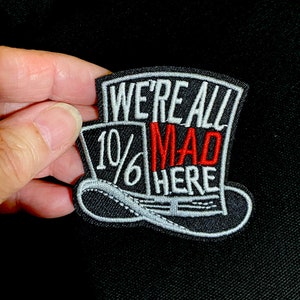 以下が含まれることがあります： 黒色の刺繍パッチ。シルクハットのデザインと「WE'RE ALL MAD HERE」の文字。数字の「10/6」も表示されています。「MAD」の文字は赤色です。
