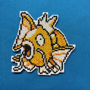 Peut inclure: Un écusson brodé au point de croix représentant un personnage de poisson pixélisé jaune et blanc avec une bouche grande ouverte, des contours noirs et des accents orange. Le motif est réalisé en points de grille sur un support en tissu blanc.