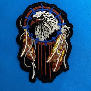 Può includere: Toppa ricamata con la testa di un'aquila dettagliata all'interno di un design acchiappasogni. La toppa presenta dettagli marroni, bianchi, blu e gialli, con piume pendenti. Ha un bordo nero ed è adatta per abbigliamento o accessori.