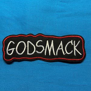 May include: A rectangular embroidered patch with a black background and a red border. The word GODSMACK is stitched in white capital letters across the center. The patch has an irregular, wavy edge following the contour of the text.