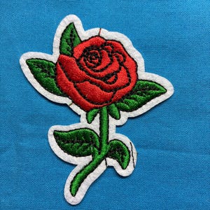 May include: An embroidered patch featuring a vibrant red rose with detailed black stitching, accompanied by green leaves and a green stem, all outlined with a white border against a solid blue background.