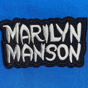 May include: A rectangular embroidered patch featuring the text Marilyn Manson in white, stylized, bold lettering. The letters are stitched onto a solid black fabric background with a scalloped, black-stitched border, set against a bright blue fabric.