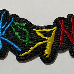 May include: A black embroidered patch with the word "KOЯN" in colorful stylized lettering. The letters are outlined in black and filled with blue, green, yellow, and red thread.