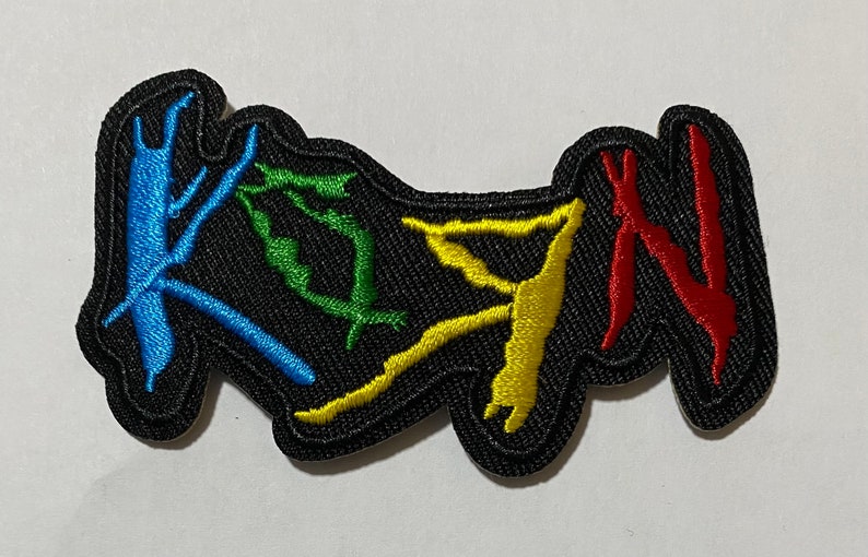 May include: A black embroidered patch with the word "KOЯN" in colorful stylized lettering. The letters are outlined in black and filled with blue, green, yellow, and red thread.