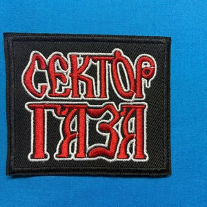 Könnte beinhalten: Schwarzer rechteckiger Aufnäher mit dem kyrillischen Text "CEKTOP ГAЗA" in Rot mit weißem Umriss. Der Aufnäher befindet sich auf einem blauen Hintergrund.