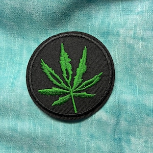 Puede incluir: Un parche redondo bordado en negro con un diseño de hoja de cannabis verde. La hoja está centrada en el parche, con costuras detalladas. El parche está sobre un fondo azul claro texturizado.