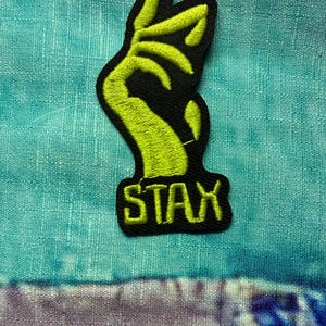 May include: A black and neon green embroidered patch featuring a hand gesture with the word "STAX" in a block font. The patch is on a teal and purple patterned fabric.