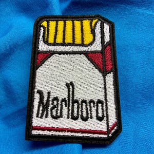 May include: A rectangular embroidered patch featuring a graphic of a cigarette pack with a white background, red accents, and yellow top details. The word Marlboro is stitched in black lettering across the center, all outlined in a thick black border.