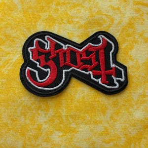 Könnte beinhalten: Gestickter Aufnäher mit dem Wort "GHOST" in gotischer Schrift. Der Aufnäher hat einen schwarzen Hintergrund mit roter und weißer Stickerei, vor einem gelben Stoff mit Muster.